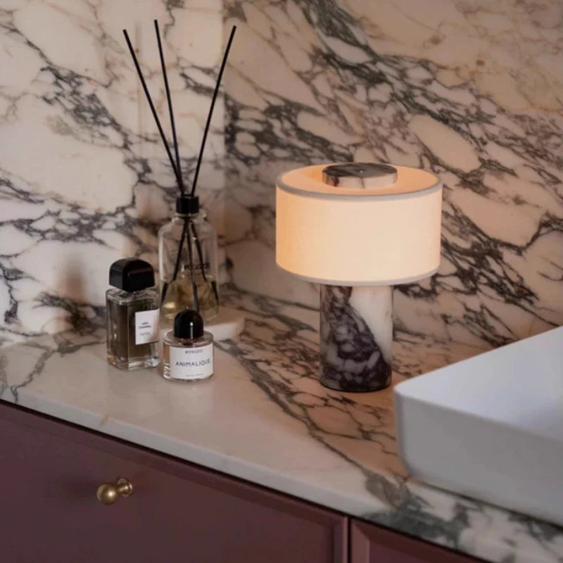 Lampe Isaure allumée dans une salle de bain en marbre, avec flacons et diffuseur de parfum