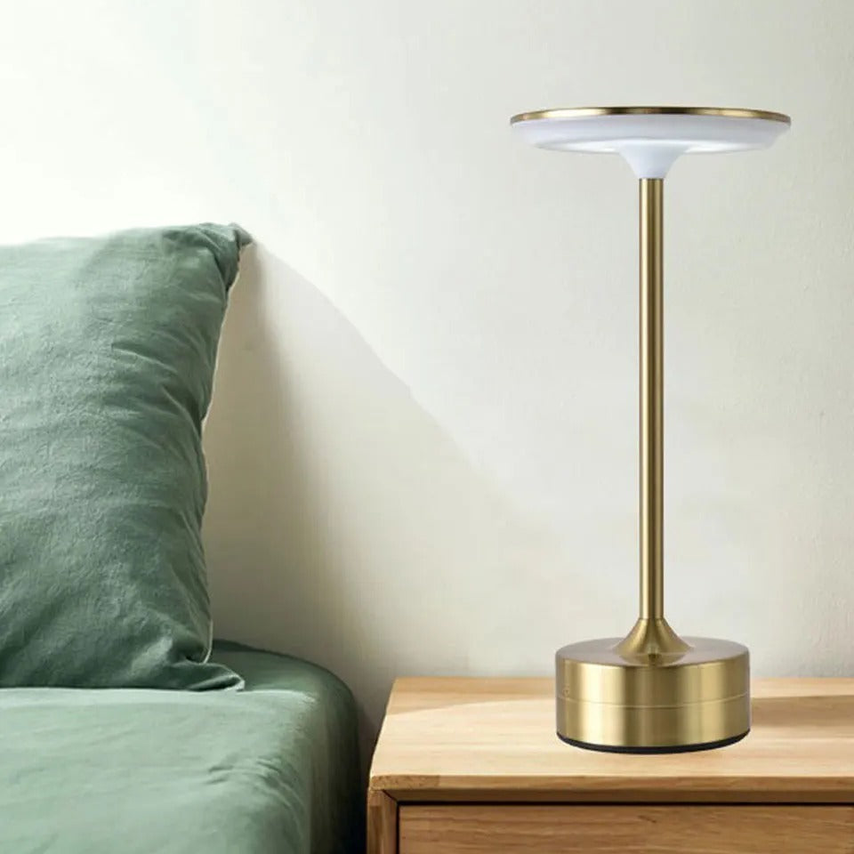 Lampe Domitille dorée sur une table de chevet en bois à côté d'un lit au linge vert